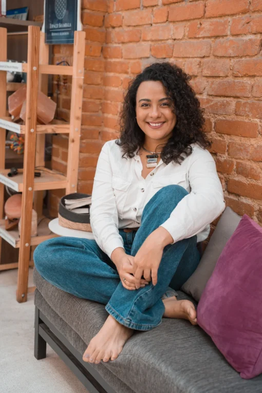 Débora Lucas sentada em um sofá, sorrindo, em um ambiente acolhedor, transmitindo leveza e propósito.